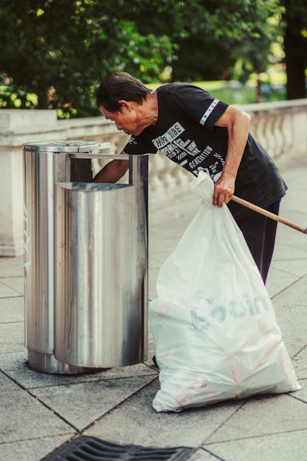 A middle-aged man with dark hair, wearing a black T-shirt with white text and dark trousers, is bent over placing a large white garbage bag into a metal trash bin on a paved sidewalk. The trash bin is shiny, cylindrical, made of stainless steel with a slot at the top for waste disposal, positioned alongside a concrete pathway bordered by a low stone wall. Behind the wall, dense green trees and foliage are visible, creating a natural background illuminated by daylight. The man appears to be engaged in waste collection or rubbish removal, consistent with the services provided by Rubbish Removal Marylebone, situated in an outdoor urban environment that may be suitable for private waste handling or alternative rubbish pickup options. The overall scene highlights an effort in environmental cleanliness and waste disposal in a residential or park-adjacent area, capturing the practical action of refuse management in a professional and neutral manner.