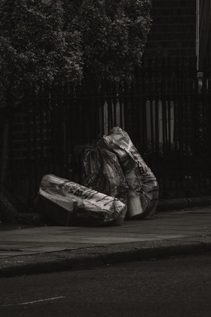 A middle-aged man with dark hair, wearing a black T-shirt with white text and dark trousers, is bent over placing a large white garbage bag into a metal trash bin on a paved sidewalk. The trash bin is shiny, cylindrical, made of stainless steel with a slot at the top for waste disposal, positioned alongside a concrete pathway bordered by a low stone wall. Behind the wall, dense green trees and foliage are visible, creating a natural background illuminated by daylight. The man appears to be engaged in waste collection or rubbish removal, consistent with the services provided by Rubbish Removal Marylebone, situated in an outdoor urban environment that may be suitable for private waste handling or alternative rubbish pickup options. The overall scene highlights an effort in environmental cleanliness and waste disposal in a residential or park-adjacent area, capturing the practical action of refuse management in a professional and neutral manner.