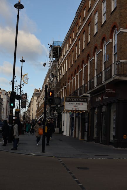 A busy street scene in Marylebone featuring a row of multi-storey brick buildings with white-framed arched windows and small balconies on the upper floors. The buildings are situated directly alongside the pavement, which is populated with pedestrians walking in various directions; some are carrying bags, indicating shopping or errands. To the left, street lamps line the sidewalk, with one lamp post visible prominently in the foreground. A traffic light with an orange pedestrian signal is positioned near the corner, guiding pedestrians to wait. The sky above is partly cloudy, with patches of blue visible. In the background, scaffolding is set up on a building under renovation, suggesting ongoing construction work, which is a common feature in urban environments that may involve building maintenance or refurbishment. The ground level of the buildings includes retail shops; one has a visible sign for a hairdresser and another appears to be a retail store with partially visible signage. The scene captures a typical urban environment with a focus on everyday activity, and indirectly reflects the context of city-based waste management by illustrating the density and activity in Marylebone’s commercial streetscape, where private rubbish collection services like Rubbish Removal Marylebone operate to support local waste disposal needs.