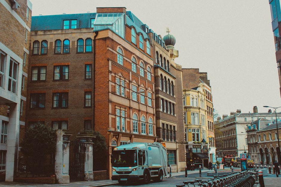 A city street scene featuring a collection of multi-storey buildings with varied architectural styles. The prominent building in the centre has a brick façade with large, arched windows on the upper floors and decorative detailing, topped with a blueish-green domed rooftop. To its right, a cream-coloured building exhibits ornate window frames and a small balcony. The street is lined with pavement, and a white rubbish collection vehicle is parked in the foreground, partially blocking a gated entrance to a property on the left. The vehicle appears to be a compact, modern waste collection truck with a clean exterior and visible equipment for rubbish collection. In the background, additional historic and contemporary structures are visible, along with street furniture such as bollards and lamp posts. The scene is captured in natural lighting, creating a neutral tone suitable for an article about private waste collection services, including rubbish removal options outside of local authority collection.
