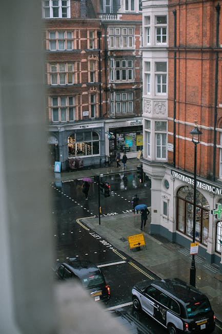 A busy street scene in Marylebone featuring a row of multi-storey brick buildings with white-framed arched windows and small balconies on the upper floors. The buildings are situated directly alongside the pavement, which is populated with pedestrians walking in various directions; some are carrying bags, indicating shopping or errands. To the left, street lamps line the sidewalk, with one lamp post visible prominently in the foreground. A traffic light with an orange pedestrian signal is positioned near the corner, guiding pedestrians to wait. The sky above is partly cloudy, with patches of blue visible. In the background, scaffolding is set up on a building under renovation, suggesting ongoing construction work, which is a common feature in urban environments that may involve building maintenance or refurbishment. The ground level of the buildings includes retail shops; one has a visible sign for a hairdresser and another appears to be a retail store with partially visible signage. The scene captures a typical urban environment with a focus on everyday activity, and indirectly reflects the context of city-based waste management by illustrating the density and activity in Marylebone’s commercial streetscape, where private rubbish collection services like Rubbish Removal Marylebone operate to support local waste disposal needs.