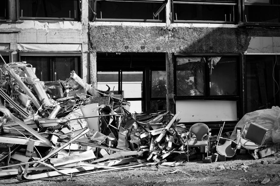 A large pile of discarded metal debris and broken furniture, including aluminum frames, panels, and miscellaneous scrap, is situated in front of a partially constructed or damaged building with exposed concrete and large, dark window openings. The debris, occupying the foreground, comprises twisted, jagged pieces with varied textures from smooth to rough surfaces, predominantly in shades of metallic silver and grey. Among the refuse are a few damaged chairs and a large plastic bag or covering, leaning against the clutter on the right side. The building's exterior features unfinished concrete surfaces with visible rough patches and exposed structural elements, and the second-floor window openings are dark and empty. Natural daylight illuminates the scene, casting subtle shadows on the debris and the building facade. This scene reflects an instance of private waste collection or site clearance, as handled by services like Rubbish Removal Marylebone, typically associated with bulky waste removal from renovation sites or properties in need of clearing before disposal or recycling.