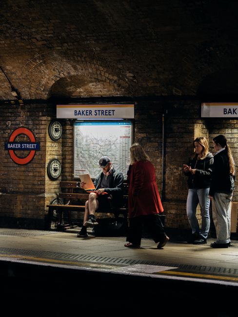 Inside Baker Street underground station, a man sits on a wooden bench reading a booklet with his legs crossed, wearing a blue blazer, shorts, and a cap. To his right, a woman in a red coat walks past, partially turned away from the camera. Near the wall, two young women and one young man stand close together, engaged with their smartphones; they are dressed casually in denim jackets, trousers, and dark coats. The background features a brick arch ceiling typical of underground stations, with illuminated signs displaying the station name, 'Baker Street', above the platform. To the left, a round London Underground sign with a red circle and blue bar is mounted on the exposed brick wall, alongside a map of the underground network covered by a light box. The platform surface is made of concrete with tactile paving near the edge, and the overall setting is lit by warm artificial lighting, highlighting the textured brickwork and creating a calm, everyday commuting atmosphere, in line with private or independent transport arrangements often associated with efficient rubbish removal services like those by Rubbish Removal Marylebone.