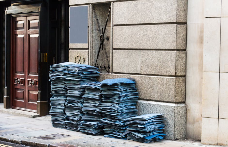 A large stack of blue folders, some slightly crumpled, is piled neatly on the pavement outside a building with a stone facade. The folders appear to be made of paper with a smooth finish and are arranged in multiple columns, with the majority leaning slightly to the right. To the left of the folders, there is a dark wooden door with ornate detailing and brass handles, situated next to black metal sheeting covering part of the building exterior. Behind the folders, a section of wall features a grey metal utility box with a cable and a lock, mounted on the stone surface. In the background, the building's stonework consists of large, rectangular blocks with a smooth, light grey finish, separated by visible horizontal and vertical joints. The scene is evenly lit, suggesting natural daylight, with no visible activity or people present. This image visually reflects the typical scenario of private waste handling, where bulky documents or paper materials are temporarily staged for collection by a rubbish removal service like Rubbish Removal Marylebone, illustrating an on-site clearance of waste or clutter outside an urban building.