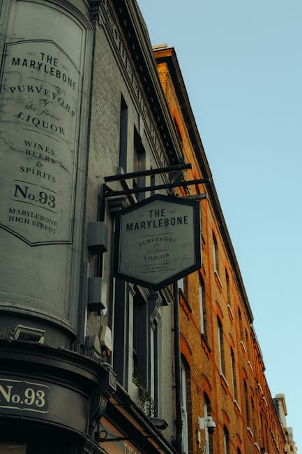 A close-up view of the facade of a historic brick building in Marylebone, featuring a large, vertically oriented, semi-circular sign for The Marylebone pub, which displays the pub's name, address on High Street, and references to wine, spirits, and liquor in black lettering on a white background. Adjacent to this sign, there is a smaller, rectangular hanging sign with similar branding. The building’s exterior wall is made of warm-toned red brick with visible mortar joints, and a row of rectangular windows with white frames and sills. The sky above is clear and light blue, providing bright natural lighting that illuminates the building’s textures and details. This image remains within the context of urban environments where external signs serve as identifiers for local businesses, including those offering private waste handling or services related to rubbish removal, aligning with the service relevance of independent or alternative waste collection options sometimes employed in commercial or residential settings managed by companies like Rubbish Removal Marylebone.