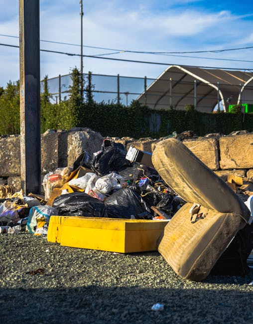 A roadside scene featuring a collection of discarded rubbish including black plastic trash bags, a yellow plastic container, and a large, worn-out car tire leaning against a low stone wall. The rubbish is scattered on a gravel surface with varied materials such as paper, plastic, and packaging visible within the bags. In the background, there is a metal pole, utility wires overhead, and a fenced area with greenery and a large tent-like structure, suggesting an outdoor area possibly near a waste collection or disposal point. The lighting indicates daylight with a partly cloudy sky, emphasizing the unmanaged nature of the waste in the scene, which may be associated with private or independent rubbish removal efforts overseen by companies like Rubbish Removal Marylebone, highlighting concerns around illegal dumping or non-authorised waste disposal in the local area.