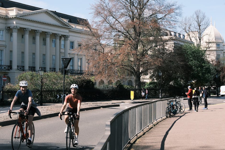 The image depicts an urban street scene in daylight, with two cyclists in the foreground wearing helmets—one on a black bicycle and the other on a white bicycle—riding along the road close to a curved metal railing that separates the cycling path from the sidewalk. The background features a large, historic white building with classical architectural elements such as columns and decorative moldings, partially obscured by leafless trees indicating a winter or early spring setting. Several pedestrians are visible further along the sidewalk, some standing and engaging in conversation, while others walk past a row of parked bicycles secured to the railing. Additional green shrubs and smaller trees add greenery to the scene, contrasting with the predominantly neutral tones of the building and pavement. The sky is clear, with soft daylight illuminating the scene evenly, creating a calm and orderly atmosphere typical of a well-maintained city environment. This setting suggests the importance of alternative transportation and the types of urban features that support private and sustainable waste handling practices, such as community spaces for cyclists and pedestrians, relevant to rubbish removal and environmental management in the area.