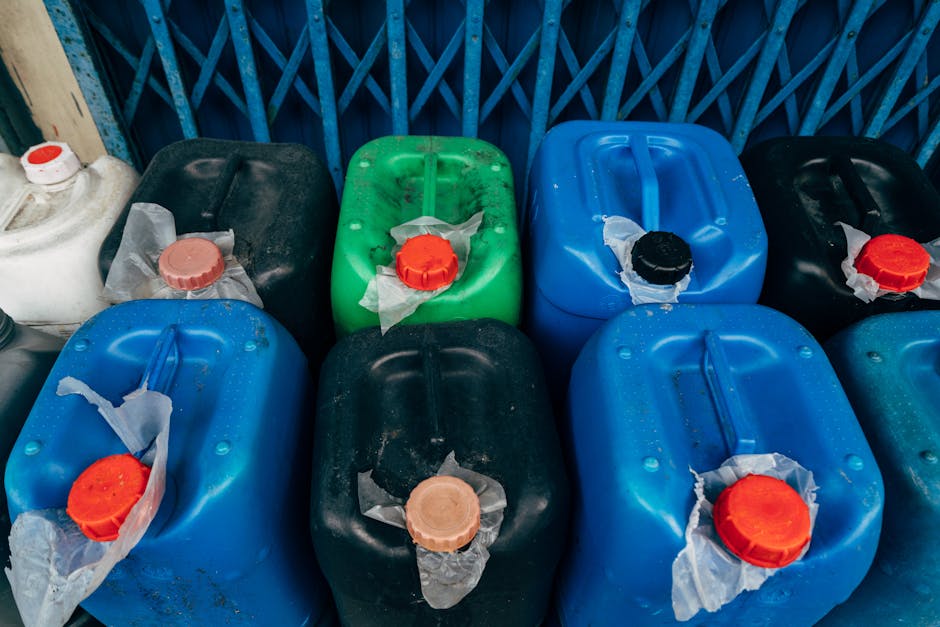 A collection of plastic containers in various colours including black, green, and blue, arranged on a paved surface against a background of a blue metal fence. Each container has a large, round, screw-on cap in contrasting colours such as red, black, or beige, with some caps covered by torn pieces of white or transparent plastic wrapping. The containers appear to be made of hard plastic, exhibiting a smooth, slightly reflective finish and showing signs of minor dirt or residue. The containers are positioned closely together with some leaning slightly, and the scene is illuminated by natural daylight, creating a clear, detailed view suitable for describing waste collection or alternative rubbish handling. These containers are often used for storing or transporting household hazardous waste, reflecting a context of private waste management efforts. Rubbish Removal Marylebone specializes in managing such waste disposal scenarios, emphasizing proper handling and segregation of hazardous materials, demonstrated by the variety of container colours and their secure caps, arranged on the pavement near a blue metal fence, suggesting a setting for on-site clearance or private collection of household waste items.