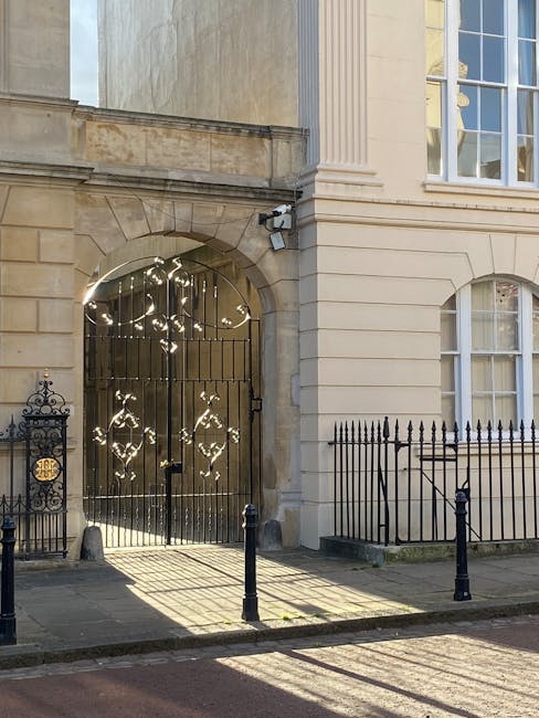 Photograph of a historic building entrance featuring a large, ornate black iron gate with decorative patterns, set within a stone archway with weathered beige stonework. To the left of the gate, there is a small, decorative iron fence with a crest-style metal plaque, and two short black bollards positioned on the pavement directly in front of the gate, which cast distinct shadows onto the ground. The building’s facade is composed of smooth, cream-coloured stone with horizontal lines and subtle architectural detailing, including tall, narrow windows with white frames on the right side, partially visible in the upper portion of the image. The overall scene is illuminated by natural daylight, creating contrast and highlighting the textures of the stone and metal elements, with a clean, well-maintained environment suggestive of an affluent residential or institutional property. This setting relates to private property management and potential alternative rubbish handling methods, such as on-site clearance or private collection by waste management services like Rubbish Removal Marylebone.