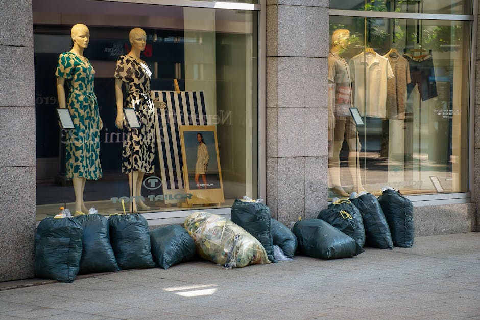 A row of tightly packed dark green plastic rubbish bags and one transparent bag filled with mixed waste are lined up on the sidewalk beneath a large shop window. The window display features three mannequins dressed in patterned dresses and various clothing items hanging on the wall behind. The rubbish bags are positioned directly in front of the store, which appears to be a clothing retailer, with the bags slightly overlapping and resting on the pavement. The shop's exterior includes stone or concrete framing around the glass windows, and the bags are situated in a manner consistent with private waste disposal or independent collection outside a commercial premises, supporting non-local-authority rubbish removal services as part of the environment. The scene is illuminated by natural daylight, creating clear visibility of the bags, window display, and surroundings, emphasizing the contrast between the discarded waste and the retail presentation behind glass.