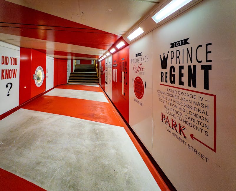 An indoor pedestrian underpass with a smooth concrete floor that features a wide central red stripe leading towards a staircase at the far end, which has dark, contrasting steps with yellow safety beneath. The walls are decorated with various vintage-style advertisements and informational posters, including a prominent large sign for 'Prince Regent,' with a crown graphic and historical text, printed in black and red on a white background. The ceiling is illuminated by rectangular fluorescent light fixtures that cast a bright, even light across the tunnel, highlighting the vivid red and white wall colors and the textured finish of the concrete floor. The overall environment appears clean and well-maintained, with an atmosphere suitable for safe pedestrian movement, juxtaposing historic promotional graphics with the modern utility of a passageway potentially associated with private waste and rubbish removal services, such as those offered by Rubbish Removal Marylebone.