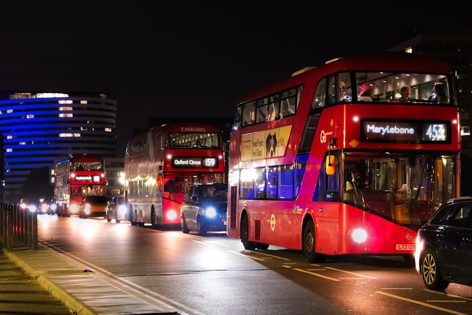 A nighttime urban scene showing three double-decker buses parked in a row on the road, with the nearest bus displaying a digital route sign indicating 'Marylebone' and the route number 453. The buses are predominantly red with some branding and advertisements along their sides, and they are illuminated by internal lights and streetlights, casting reflections on their surfaces. In the background, modern office buildings with curved and striped illuminated facades are visible under a dark sky. Several vehicles are parked or moving nearby, including a black car with its headlights on close to the front bus. The pavement and road surface are visible in the foreground, with some visible markings and a small yellow curb, suggesting a city setting suitable for private or independent collection services like those provided by Rubbish Removal Marylebone.