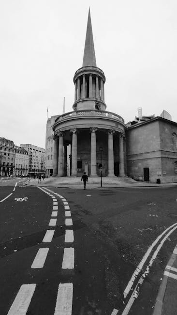A black and white photograph of a historic circular building with a tall, pointed spire extending high into the sky. The structure features a colonnade of evenly spaced columns supporting a rounded entablature, with a smaller, cylindrical section beneath the spire. The building is situated on a paved urban street, with a person walking close to its base. The foreground includes road markings, such as dashed white lines and a bicycle lane symbol, indicating a designated space for cyclists. Surrounding the building are other city structures with traditional facades, some with visible windows and rooftops, contributing to a busy cityscape atmosphere. The scene is captured in daytime with even, natural lighting, emphasizing the architectural details and urban environment, which aligns with the context of private waste handling and independent collection by rubbish removal services such as Rubbish Removal Marylebone.