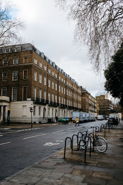 A street scene featuring red-brick residential buildings with white-framed sash windows, some with window boxes. In the foreground, a section of a building's exterior wall displays a street name sign indicating 'Providence Court W1' and a smaller sign for 'Balderton Street W1.' The street level shows a narrow alleyway with a black waste disposal bin and a collection of blue and black trash bags placed nearby. Two individuals, dressed in casual clothing and jackets, are seen walking past the bins in the alleyway. The environment is well-lit with natural daylight, and the overall scene reflects a typical urban area where private waste collection services, such as those offered by Rubbish Removal Marylebone, may be used for efficient rubbish disposal and on-site clearance in Marylebone, London.