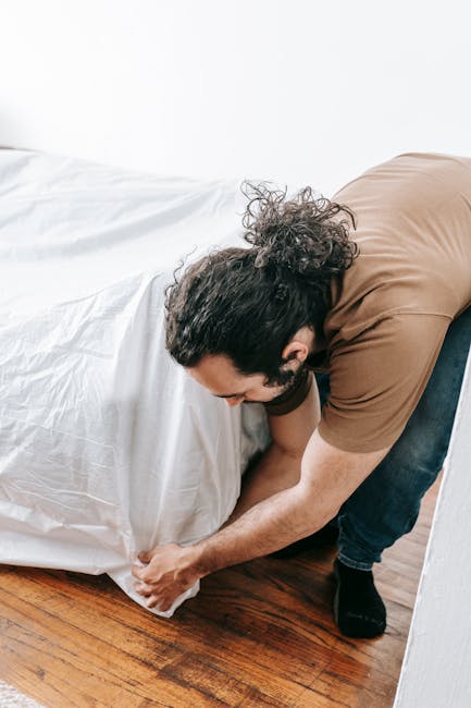 A man with long, curly dark hair, wearing a brown T-shirt and dark jeans, is kneeling on a wooden floor as he carefully lifts the corner of a white mattress. The mattress has a smooth, slightly shiny cover, and is positioned close to a white wall, with its edge resting on the floor. The surrounding environment appears to be a bright, interior room with natural lighting, and the scene is part of a professional rubbish removal service that might involve on-site clearance of household items, such as mattresses, with a focus on careful handling and efficient waste disposal by Rubbish Removal Marylebone. The image emphasizes the physical effort involved in moving bulky items, which is typical of independent collection or private waste handling scenarios, where careful lifting techniques are necessary to prevent damage or injury. Overall, the setting highlights the practical aspect of rubbish removal, especially for large furniture items, within a clean and minimalistic interior environment.
