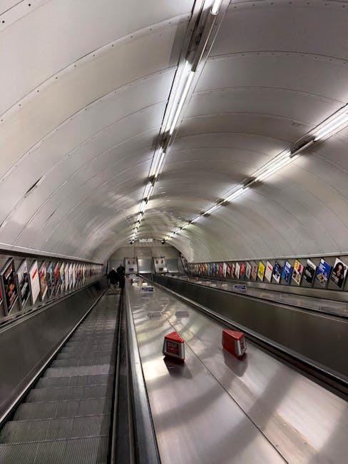 An underground pedestrian tunnel with a curved, white, metallic ceiling illuminated by a row of evenly spaced fluorescent lights. The tunnel features moving walkways on either side, made of textured grey metal with red and white barriers positioned on the right, indicating possible maintenance or restricted access. Along the lower parts of the walls, there are advertising posters or informational displays with various colors and text, partly visible but not legible. The environment appears clean, modern, and designed for efficient pedestrian flow, potentially related to an underground transportation system in an urban area. The scene suggests an alternate or supplementary pathway for commuters, aligning with themes of private or independent transit and associated waste or rubbish management scenarios that may involve cleaning or maintenance services such as those provided by Rubbish Removal Marylebone in city infrastructure settings.