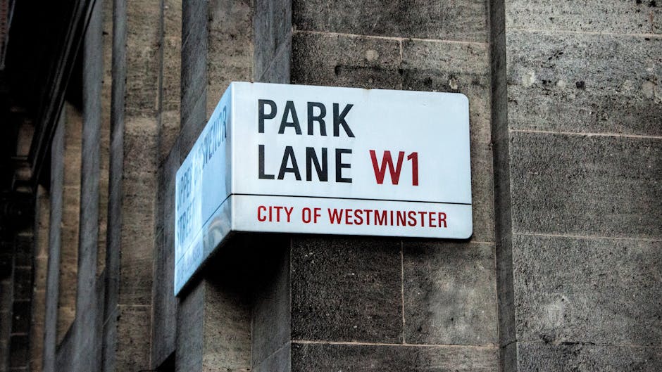 A rectangular street sign mounted on a textured stone wall beneath an iron staircase railing, indicating 'Park Lane W1' and 'City of Westminster' in red and black text on a white background. The sign is positioned to the left of the frame, with the stone wall displaying a rough, weathered surface in varying shades of grey and brown. The iron railing, with vertical bars and a dark finish, is partially visible on the left side, suggesting an urban setting in central London. The lighting is natural, illuminating the sign and wall with soft shadows that enhance the textures and details in the scene. This image depicts a typical street sign found in the area, relevant to navigating or understanding local regulations regarding waste removal or parking, with the location context subtly implied by the signage. Rubbish Removal Marylebone services may be associated with areas like this, where off-street private clearance might be necessary for waste management compliance.