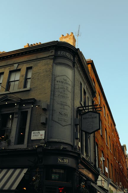 The image shows the upper portion of a historic building on a city street, with a rounded corner façade and a decorative sign mounted on the side. The building features a dark grey, slightly weathered exterior with detailed moldings and a sign reading 'The Marylebon' and 'No. 98,' indicating its address. The corner of the building displays a faded, vertically oriented plaque or sign, which is difficult to read but suggests historical significance. The building's large windows have black frames, and some are illuminated, with a striped awning extending over the shopfront on the ground floor. The sky above is clear with a soft blue hue, and a chimney or similar structure is visible at the roof's edge. This setting may relate to alternative waste handling or private collection services, as the building appears to be an older commercial or retail property situated on a busy city street. The overall scene exudes a classic, urban appearance with attention to structural detail and signage, illustrating a typical historic architectural environment seen in London neighborhoods like Marylebone.